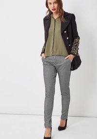 Blazer negro con botones dorados y mangas con estampado floral, top verde oliva y pantalones de cuadros blanco y negro. Tacones altos negros.