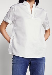 Vit bomullstopp med korta ärmar, ståkrage och knappslå. Knappar i sidan och en ledig passform, kombinerad med blå jeans.