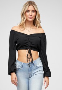 Fekete, váll alatti crop top, hosszú, puffos ujjakkal, ráncolt elülső résszel és állítható kötővel. Világoskék farmernadrággal párosítva.