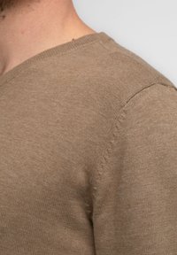Bruine gebreide trui met een V-hals, voorzien van een gladde textuur en subtiele naaddetails op de schouder. Standaard pasvorm.