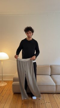 Grijze joggingbroek met witte elastische tailleband, vastgehouden door een persoon in een zwart longsleeve shirt, tegen een beige bank en houten vloer.