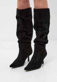 Botas de ante negras sobre la rodilla con puntera apuntada y tacón alto delgado. Cuenta con una textura recogida en la parte superior para un efecto arrugado.