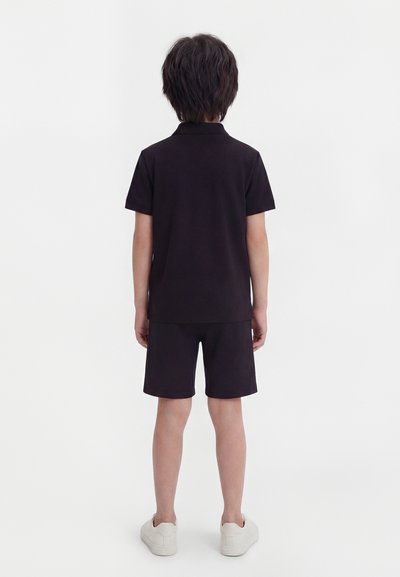 Παιδί που στέκεται γυρισμένο προς τα πίσω, φορώντας μαύρο πόλο μπλουζάκι με κοντά μανίκια, μαύρο σορτς και λευκά αθλητικά παπούτσια, μπροστά σε απλό λευκό φόντο.