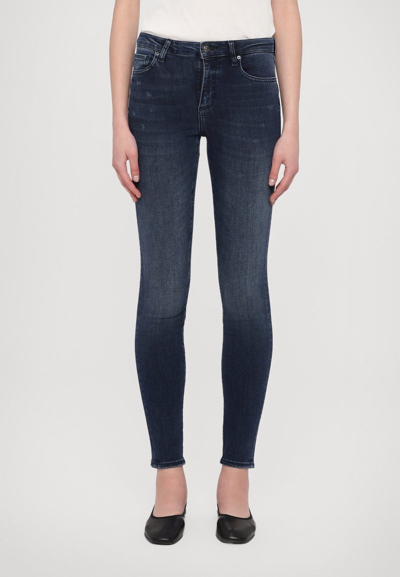 Femme portant un jean skinny bleu foncé et des ballerines noires, debout, les bras détendus le long du corps, devant un fond uni.