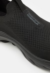 Zapato negro Skechers Go Walk con parte superior de malla, diseño sin cordones y superficie texturizada, con suela de goma flexible y detalle del logo.