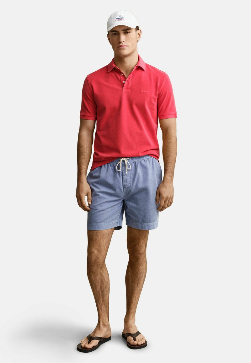 Junger Mann trägt ein rotes Poloshirt, hellblaue Shorts mit Kordelzug, weiße Mütze und schwarze Flip-Flops, steht vor einem einfarbigen Hintergrund.
