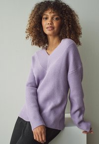 Maglione a maniche lunghe con scollo a V di colore viola chiaro, con texture a coste. Il modello lo ha abbinato a un pantalone scuro a righe sottili, su uno sfondo neutro.