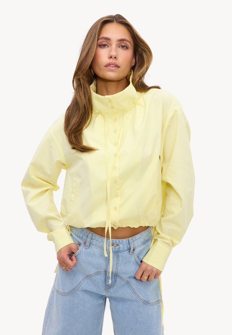 Femme aux longs cheveux portant une veste jaune à col montant boutonnée et un jean bleu clair, les mains dans les poches, sur fond blanc.