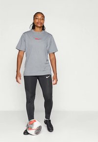 Szara sportowa koszulka z czerwonym logo, czarne legginsy z teksturą i logo Nike oraz czarne korki piłkarskie. Przy stopach znajduje się piłka nożna.