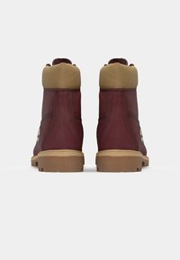 Botas de tobillo burdeos con un cuello marrón claro, suela de goma y costuras visibles. Presenta un logo en el lado exterior. Material de cuero texturizado.