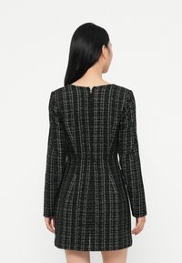Rochie neagră din material texturat, cu mâneci lungi, având un model discret în carouri, o siluetă ajustată și un detaliu de decolteu în spate sub formă de cheie.