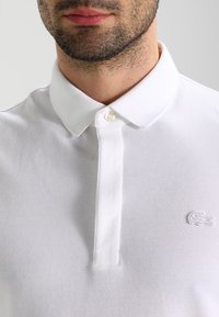 Polo shirt blanc en tissu texturé. Présente une patte de boutonnage et un petit logo brodé sur la poitrine gauche.