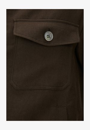 Dunkelbraune Stofftasche mit Klappe, gesichert durch einen einzelnen durchsichtigen Vierloch-Knopf, detaillierte Nähte an den Taschenrändern sichtbar.