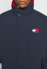 Marineblauwe jas met een hoge kraag, rode voering aan de binnenkant en een Tommy Jeans logopatch. Gestructureerde stof met zijzakken met rits.