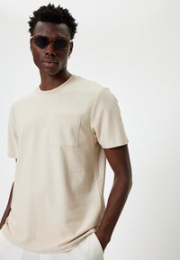 Beige kortærmet t-shirt i et blødt materiale, med en brystlomme og rund halsudskæring. Båret med runde solbriller.