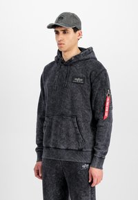 Black hooded sweatshirt with a textured finish, front pocket, and logo patch. Accompanied by a gray baseball cap. Zippered sleeve detail.