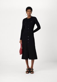 Fekete bordázott midi ruha hosszú ujjakkal, gombos elülső résszel, két mellzsebbel és fodros szegéllyel. Fekete szandálokkal és piros táskával párosítva.