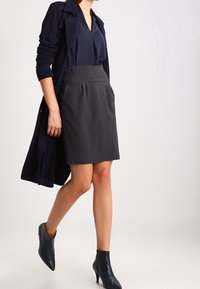 Cappotto lungo navy su una blusa scura e una gonna a matita grigia; stivali neri alla caviglia completano l'outfit. Focus su un design elegante e su misura.