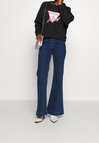 Persona con felpa nera GUESS, jeans svasati blu scuro, stivali bianchi con tacco e una piccola borsa argento con tracolla a catena.