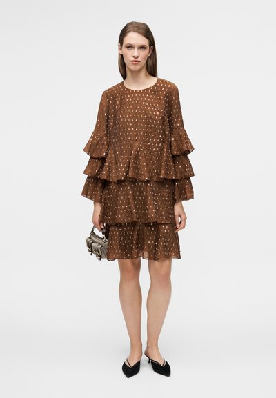 Robe marron à étages avec manches à volants et motif à pois. Tissu transparent et design en couches. Associée à un petit sac à main et des chaussures plates pointues.