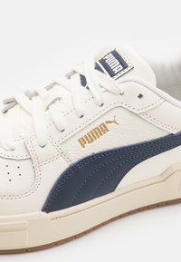 Gros plan d'une basket blanche Puma avec une bande latérale bleu marine, des lacets blancs et un logo Puma doré sur une tige en cuir texturé.