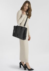 Schwarze Ledertasche mit zwei Griffen, glatter Textur und silbernen Hardware-Details, gehalten von einer Person in einem langen cremefarbenen Kleid und schwarzen Absätzen.