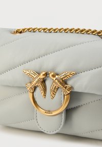 Τσάντα από ανοιχτό γκρι δέρμα με κέντημα σε σχέδιο chevron, χρυσά μεταλλικά στοιχεία με δύο πουλιά και αλυσίδα διακοσμητικό στο πάνω μέρος. Το όνομα της μάρκας "PINKO" είναι χαραγμένο.