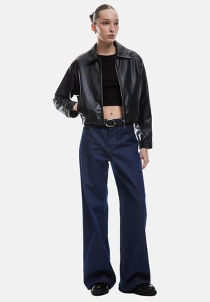 Zwart faux leren ritsjack, cropped lengte, gecombineerd met loszittende blauwe denim jeans, afgewerkt met een brede riem en zwarte schoenen.