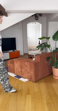 Hnedá kordurová pohovka s zaobleným tvarom, umiestnená na drevenej podlahe. V blízkosti sa nachádza zelená kvetinová nádoba, malý stolík a TV jednotka.