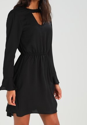 Frau trägt ein schwarzes langärmeliges Kleid mit Schlitz am Ausschnitt, elastischer Taille und ausgestellten Manschetten, steht vor einem schlichten Hintergrund.