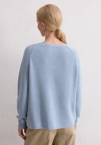 Frau mit blonden Haaren in einem tiefen Pferdeschwanz, die einen hellblauen Strickpullover und beige Hosen trägt, und vor einer weißen texturierten Wand steht.
