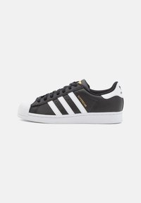 Zwart-witte Adidas Superstar sneaker met een witte shell toe, drie witte strepen, zwarte veters en "SUPERSTAR" in gouden letters aan de zijkant.
