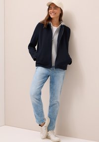 Jeune femme souriante, portant une casquette beige, une veste marine sur un sweat gris, un jean bleu clair et des baskets blanches, se tenant devant un mur beige.