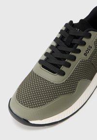 Grön nät-sneaker med svarta snören och detaljer, med en texturerad yta, rund tå och gummisula med vit rand.