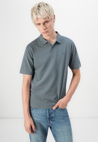 Grå kortærmet polo med krave og tre knapper, lavet af et glat stof, sat sammen med lyseblå jeans. Enkel logo detaljer.