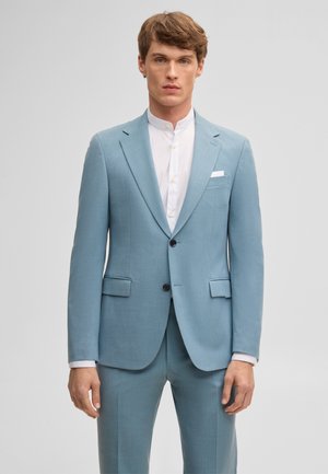 Homme portant un costume bleu clair à deux boutons avec poches à rabats et une chemise blanche col mandarin sur un fond gris uni.