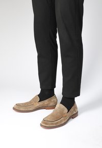 Beige Wildleder Slipper mit elastischen Seitenelementen, brauner Sohle und Nahtdetails, kombiniert mit schwarzen Hosen und Socken.