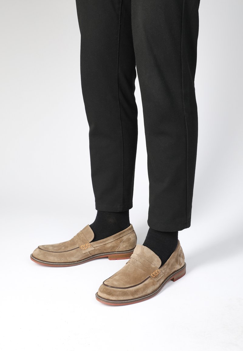Beige Wildleder Slipper mit elastischen Seitenelementen, brauner Sohle und Nahtdetails, kombiniert mit schwarzen Hosen und Socken.
