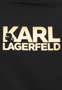 Melna T-kreklu ar izteiksmīgu zeltītu tekstu "KARL LAGERFELD" un silueta dizainu, izceļot profilu ar brillēm un astīti.