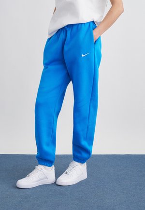 Osoba w jaskrawoniebieskich spodniach dresowych Nike i białych butach sportowych Nike stojąca na niebieskim dywanie.