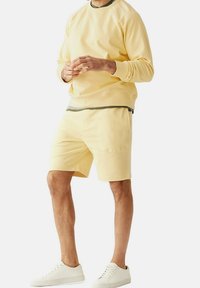 Completo maschile che include un maglione giallo chiaro con collo a coste e pantaloni corti gialli abbinati, indossato con sneakers bianche.