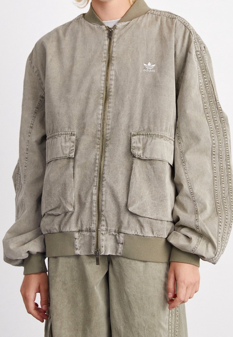 Khaki bomber jacket with a zip front, ribbed cuffs, two front pockets, and subtle adidas logo. Made from smooth fabric with a relaxed fit.