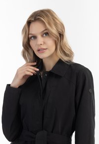 Fekete trench coat sima anyagból, galléros nyakkal, gombos záródással és oldalzsebekkel; derékon megkötős övvel.