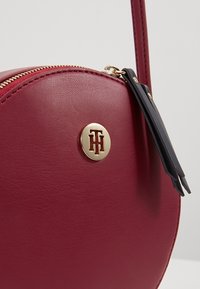 Borsa a mano rotonda in pelle bordeaux con zipper dorato e logo, dotata di una tracolla scura e texture liscia.