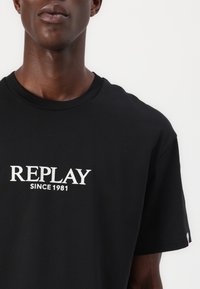 Juodas medvilnės marškinėliai trumpomis rankovėmis, su baltu užrašu "REPLAY SINCE 1981" ant priekio ir maža logotipo etikete šone.