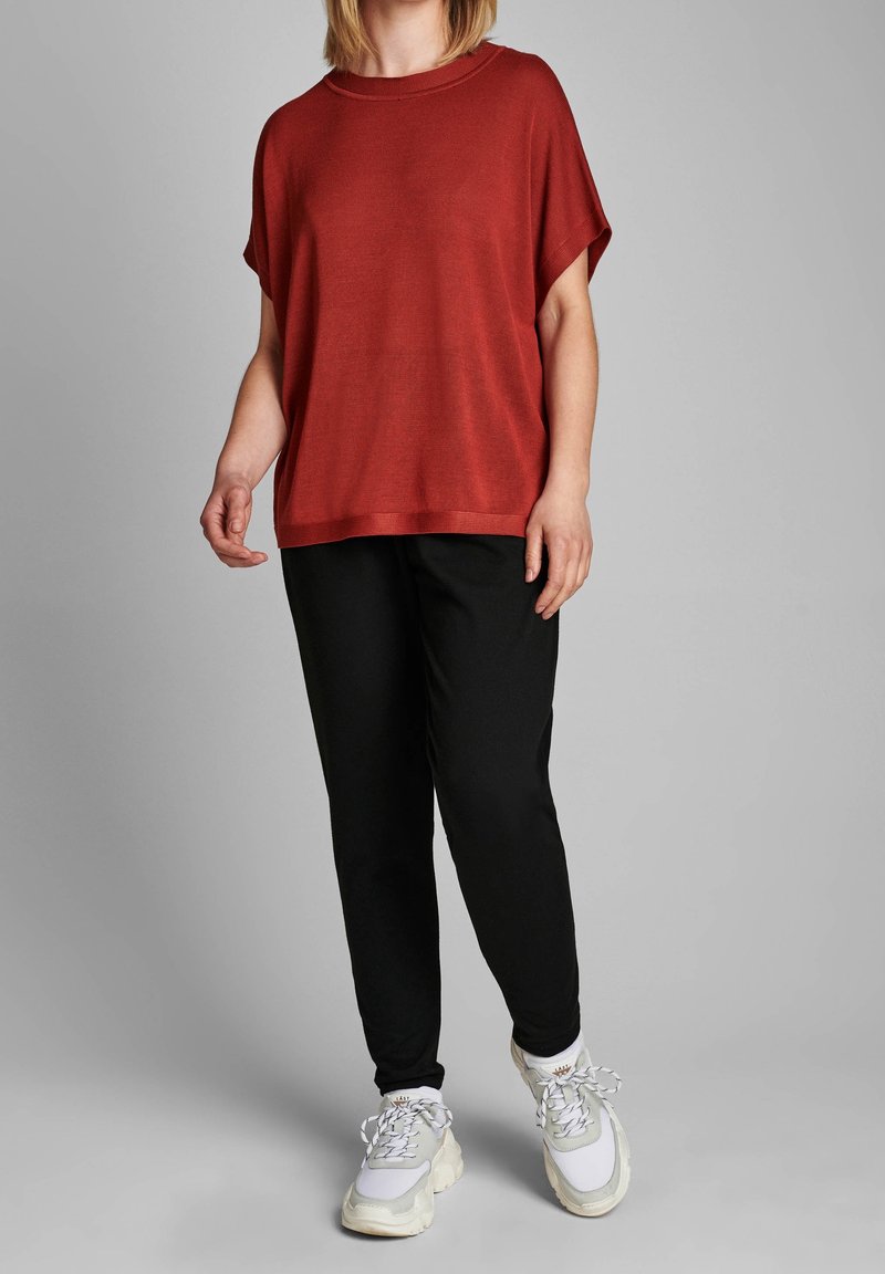 Personne portant un haut rouge à manches courtes ample, un pantalon noir et des baskets blanches épaisses, se tenant sur un fond gris.