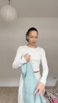 Žena se sepnutými vlasy, oblečená v bílém topu a sukni, držící světle modrý pletený oděv v minimalistické místnosti.