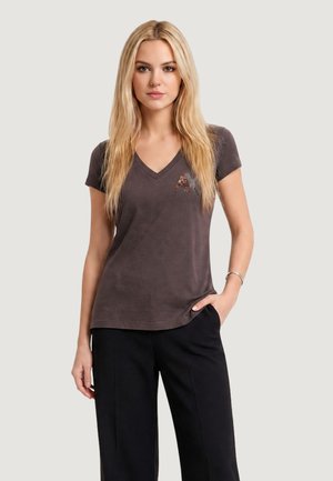 Mujer rubia vistiendo una camiseta de cuello en V gris oscuro con una letra A adornada y pantalones negros, de pie con una mano en el bolsillo.