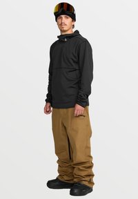 Pull noir avec une poche zippée sur la poitrine, associé à un pantalon de neige beige ample avec des zips sur les côtés. Des bottes noires et des lunettes de ski complètent le look.