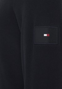 Granatowy sweter dzianinowy o teksturowanym wykończeniu i kwadratowej czarnej łatce z logo w czerwonych, białych i niebieskich akcentach na rękawie.
